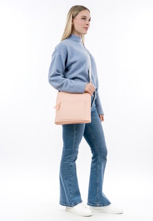 Borsa a tracolla rosa chiaro con una tasca frontale testurizzata e cerniera, abbinata a un maglione blu e jeans svasati, con scarpe da ginnastica bianche.