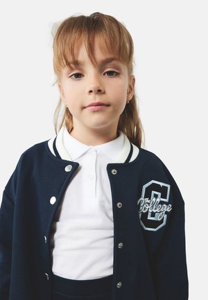 Chaqueta de varsity azul marino con cuello blanco a rayas, botones de presión y detalle bordado "La Universidad". Lleva puesta sobre una camiseta polo blanca.