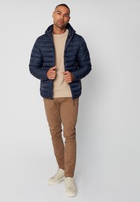 Giacca puffer blu navy con cappuccio, chiusura con zip e motivo a righe orizzontali. Indossata sopra un maglione beige e abbinata a pantaloni marrone chiaro e sneaker.