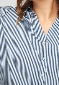 Camicia a righe blu e bianche con colletto abbottonato, caratterizzata da strisce verticali, materiale in cotone morbido e dettagli dei bottoni ben visibili.