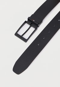 Černý kožený opasek s texturovaným povrchem, vybavený matným černým obdélníkovým přezkou a pěti otvory pro úpravu délky na pásku.