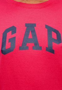 Tissu rouge avec de grandes lettres majuscules bleu foncé "GAP" imprimées sur la poitrine d'une chemise.