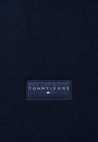 Marineblå strikket stof med en rektangulær label med "TOMMYJEANS" og et lille rød, hvid og blå horisontalt design nedenfor.