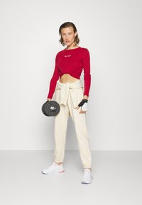 Top corto rojo de manga larga con logo, joggers beige con puños en los tobillos, zapatillas blancas, sosteniendo un disco de pesas negro, guantes sin dedos.