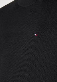Schwarzer gestrickter Pullover mit rundem Halsausschnitt und kleinem gesticktem Logo, das blaue, weiße und rote Streifen auf der Brust zeigt.