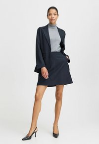 Jūrinės juostelės švarkas ir atitinkanti sijonas su pilku golfu. Modelis dėvi juodas slingback kulvines. Lygi medžiaga, pritaikytas siluetas, modernus dizainas.