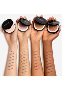 Quatre mains tiennent des étuis de maquillage compacts avec un extérieur noir. Chaque bras affiche des teintes étiquetées en texte noir, montrant une gamme de tons de peau.