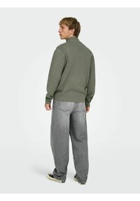 Zielony sweter z dzianiny z kołnierzykiem. Noszony z luźno dopasowanymi szarymi dżinsami i niskimi czarnymi butami.