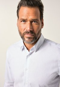 Muž s tmavými kudrnatými vlasy a vousy, oblečený v bílé košili s tenkými vertikálními pruhy, se dívá přímo do kamery.