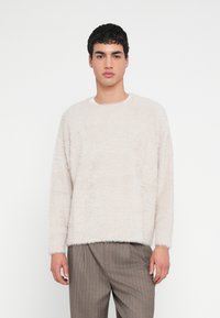 Pulover bej deschis pufos, cu decolteu rotund și mâneci lungi, având o suprafață texturată. Asortat cu pantaloni maro cu dungi.