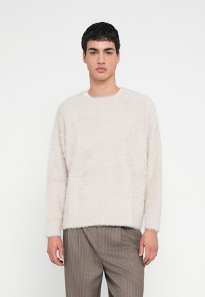 Lichtbeige, pluizige trui met een ronde hals en lange mouwen, met een textuur aan het oppervlak. Gecombineerd met gestreepte, bruine broek.