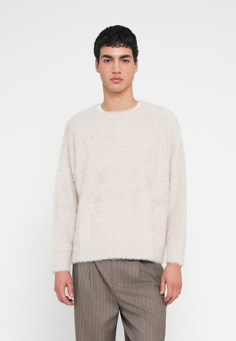 Pulover bej deschis pufos, cu decolteu rotund și mâneci lungi, având o suprafață texturată. Asortat cu pantaloni maro cu dungi.