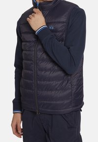 Navy puffer vest with quilted design, stand collar, front zipper, and blue accent stripes on the cuffs, worn over a long-sleeve shirt.
