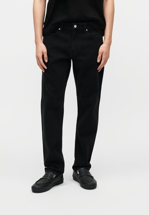 Person iført svarte jeans med rette bein, svarte slip-on sko og en svart topp med korte ermer, stående mot en ensfarget lys bakgrunn.