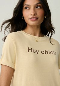 Lühikeste varrukatega, heledast kollasest pehmest kangast T-särk, millel on ees prinditud pruun tekst "Hey chick". Lihtne ja mugav disain.