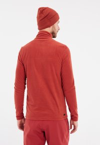 Pullover in pile rosso con colletto alto e maniche lunghe, abbinato a un berretto rosso coordinato. Tessuto morbido, branding minimale sull'orlo.
