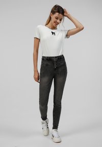 Top blanco de manga corta con ribete negro y detalle de lazo, combinado con pantalones vaqueros negros de cintura alta y zapatillas blancas. Tela suave, diseño ajustado.