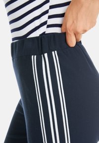 Marineblaue Jogginghosen mit einem weichen, elastischen Bund. Sie verfügen über drei vertikale weiße Streifen entlang des äußeren Beins. Das Material scheint atmungsaktiv und dehnbar zu sein.