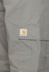 Calças cargo cinzentas em tecido com um bolso aplicado adornado com um logo da Carhartt em amarelo e roxo. O material apresenta uma textura em grade subtil.