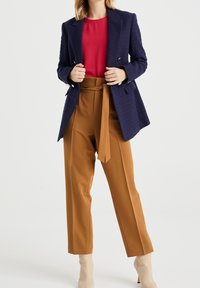 Blazer blu navy con texture e bottoni dorati, abbinato a una maglietta rossa e pantaloni a vita alta color beige. Stivali alla caviglia color crema completano il look.