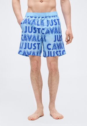 Mees, kes kannab helesiniseid ujumistrakse tumesinise abstraktse tekstuuriga, seisab paljajalu ühevärvilise valge tausta ees.