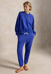 Kobieta ubrana w niebieską bluzę i spodnie dresowe ze sznurkiem w kolorze białym oraz pomarańczowe sandały, stojąca z rękami w kieszeniach na jednolitym tle.