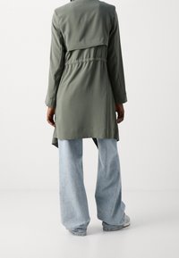 Olivgrüner Trenchcoat mit tailliertem Schnitt, langen Ärmeln und einem etwas längeren Rückenteil. Kombiniert mit hellblauen Jeans mit weitem Bein.