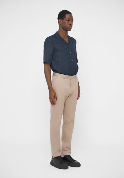 Μπλε πουκάμισο κοντό μανίκι με καπάκι και ανάγλυφο μοτίβο, συνδυασμένο με ανοιχτά μπεζ παντελόνια και μαύρα αθλητικά παπούτσια, σε λευκό φόντο.