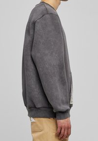 Šedá, oversized mikina s měkkou texturou a žebrovanými manžetami. Obsahuje klokánkovou kapsu a diskrétní grafický detail na boku.