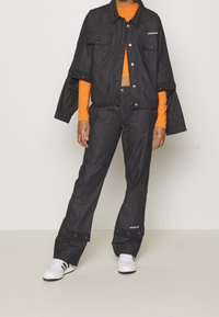 Svart denimjacka med capeslut, i kombination med svarta denimbyxor med tryckknappdetaljer, bärs över en orange topp. Vita sneakers.