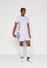 Lila Fußballuniform mit einem kurzärmligen Oberteil mit lila Akzenten und passenden Shorts. Weiße Socken und Schuhe, während ein Fußball gehalten wird.