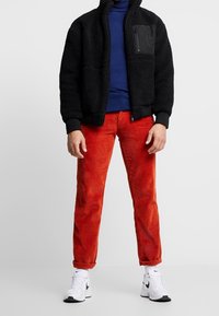 Svart fleecejacka med blixtlåsförsedd bröstficka; marinblå polotröja; röda manchesterbyxor; vita sneakers med svarta detaljer.