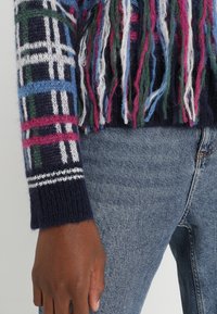 Blauer, grüner und rosa kariert Pullover mit fransigen Details, ausgestattet mit einem marineblauen gerippten Bündchen und einer gestrickten Textur, getragen über blauen Jeans.