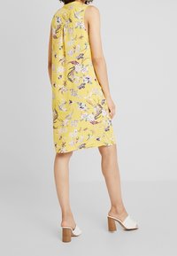 Gelbes, ärmelloses Kleid mit floralem Design und gefälteltem Rücken. Der Stoff hat eine glatte Textur mit lebendigen Blumenmustern. Weiße, Absatzmules.