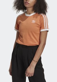 Oransjefärgad bomullströja med vit krage och ärmkanter, med tre vita ränder på varje axel och en broderad Adidas-logga.