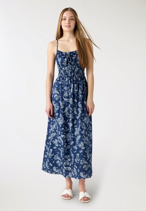 Giovane donna con capelli lunghi che indossa un vestito lungo blu senza maniche con motivi paisley bianchi e sandali bianchi a ciabatta, ferma in piedi.