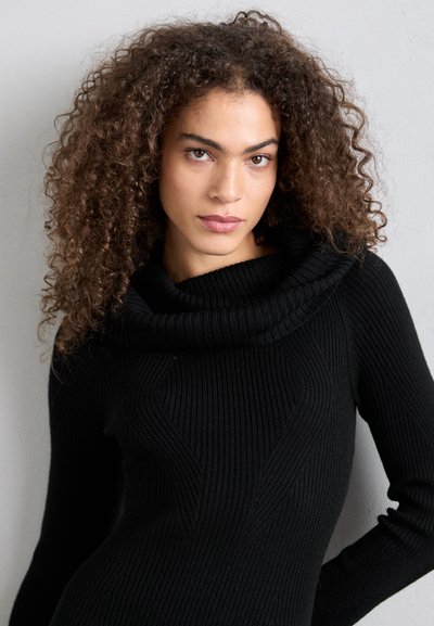 Woman with curly hair wearing a black ribbed sweater with a wide folded collar, posing against a plain light-colored wall.