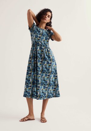 Vrouw staat blootsvoets en draagt een blauwe knie-lange jurk met patroon en korte mouwen, poseert met haar handen bij haar nek.