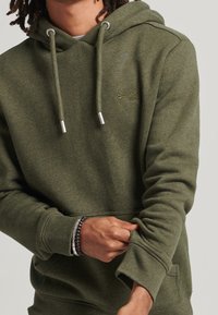 Sudadera de color verde oliva con cordones ajustables, fabricada en tejido suave, con puños de canalé y bolsillo tipo canguro. Presenta un pequeño logo en el pecho.