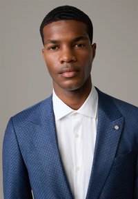Blazer blu con motivo a disegni e una texture particolare, indossato con una camicia bianca sotto e un tocco d'argento sul rever.