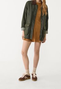 Groene katoenjack met een bruine corduroy kraag, twee voorzakken en geruite mouwen, gecombineerd met een bruine suede jurk en bruine sneakers.