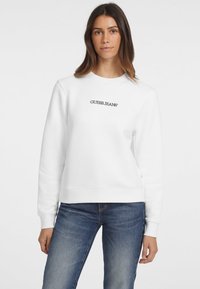 Valkoinen huppari, jossa on ribbireunat hihansuissa ja helmoissa, ja keskellä musta "GUESS JEANS" -logo. Yhdistettynä sinisiin farkkuihin.