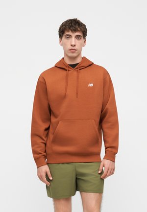 Jeune homme portant un sweat à capuche New Balance couleur rouille avec poche avant et short vert olive, debout devant un fond clair uni.