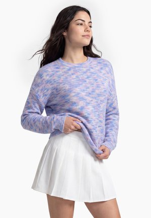 Kvinde med langt mørkt hår iført en pastel lilla og pink sweater og en hvid plisseret nederdel, der holder i sweaterens kant med den ene hånd.