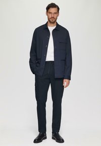 Marineblauwe jas met knopen en zakken, wit shirt, marineblauwe cargobroek en zwarte schoenen. Glad textuur, modern ontwerp.