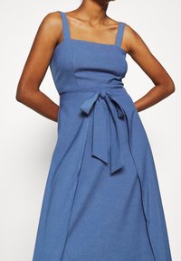 Mujer vestida con un vestido azul sin mangas, con escote cuadrado y cinturón atado en la cintura, de pie con las manos detrás de la espalda.