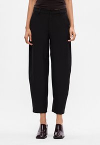 Crne široke hlače s glatkom teksturom, s ravnim pojasom i suptilnim naborima. Uparen s sjajnim tamnim cipelama.