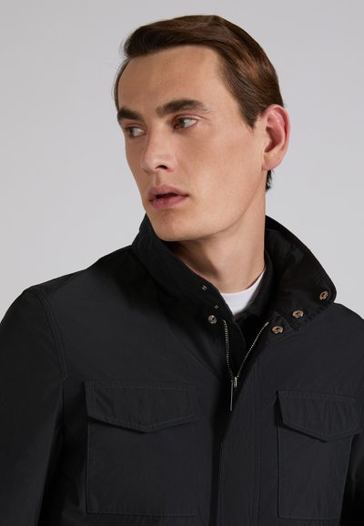 Młody mężczyzna o brązowych włosach, ubrany w czarną kurtkę na zamek z klapami na kieszeniach, patrzący na bok na neutralnym tle.