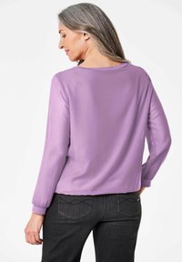 Lila Langarmbluse mit lockerer Passform und Rundhalsausschnitt. Glattes Material mit subtilem Glanz. Kombiniert mit dunklen Jeans.