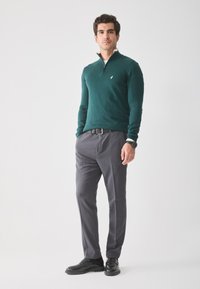 Pulover tricotat de culoare verde închis, cu guler cu jumătate de fermoar, purtat cu pantaloni gri de costume și pantofi negri. Puloverul are un mic logo pe piept.